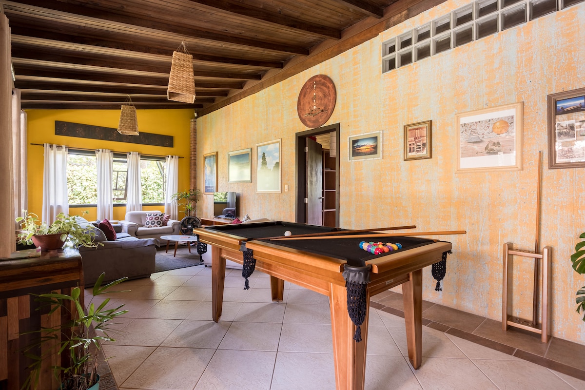 A spacious game room features a pool table positioned at the center, surrounded by warm yellow walls adorned with framed artwork. Natural light filters through large windows, illuminating a cozy seating area in the background, which includes a comfortable sofa and decorative plants.