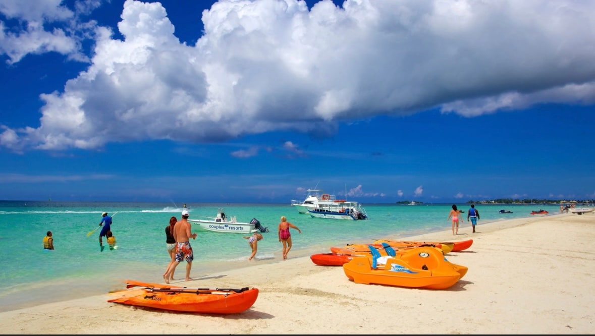 A serene beach scene showcases golden sand and clear turquoise waters. Several individuals are seen enjoying the shoreline, with a small boat approaching. Brightly colored kayaks are lined up on the sandy beach, providing access for water activities under a partly cloudy sky.