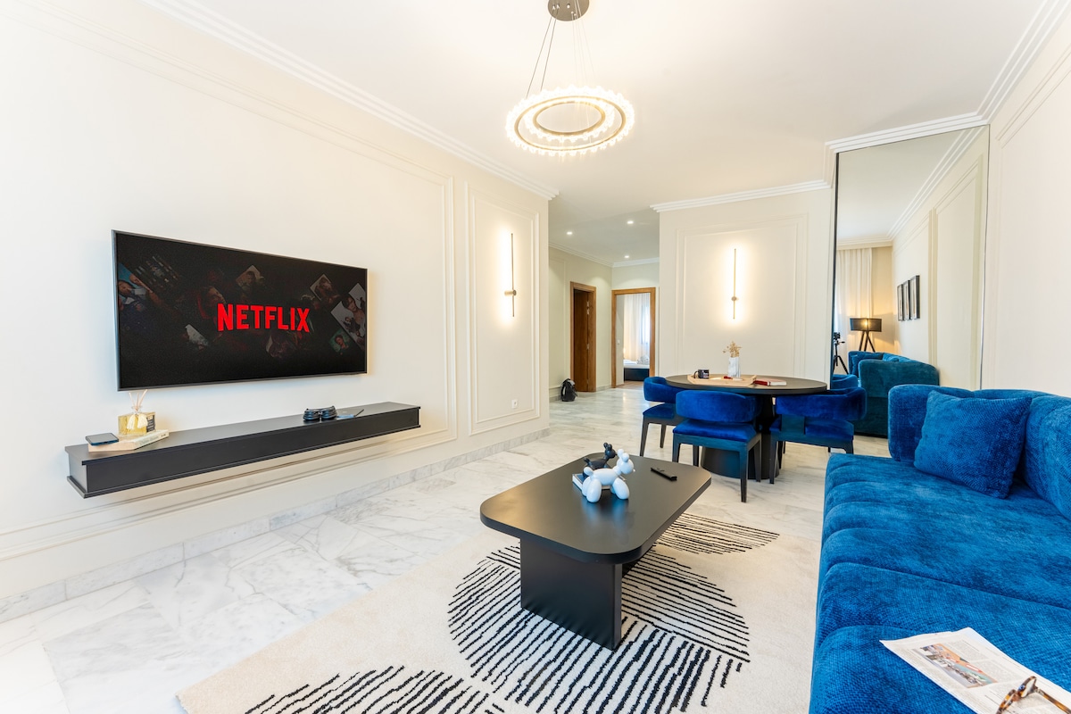 A bright living area features a blue sofa paired with dark accent chairs, creating a spacious seating arrangement. A large wall-mounted TV displays the Netflix logo. A coffee table sits atop a patterned rug, while a mirrored wall enhances the room's light and openness.