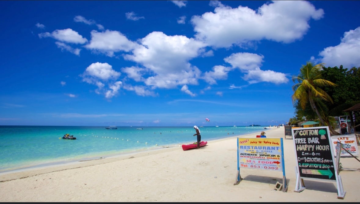A sandy beach stretches under a bright blue sky with scattered clouds. Calm turquoise water gently laps at the shore, and a paddleboarder is visible in the distance. Signs for nearby restaurants and activities are placed along the beach, offering information to visitors.