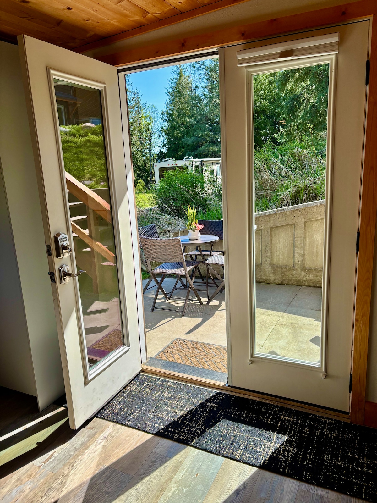 Double French doors open to a private patio area, featuring outdoor furniture including a table and chairs. Sunlight streams through the glass doors, illuminating the entryway and creating a connection to the natural surroundings.