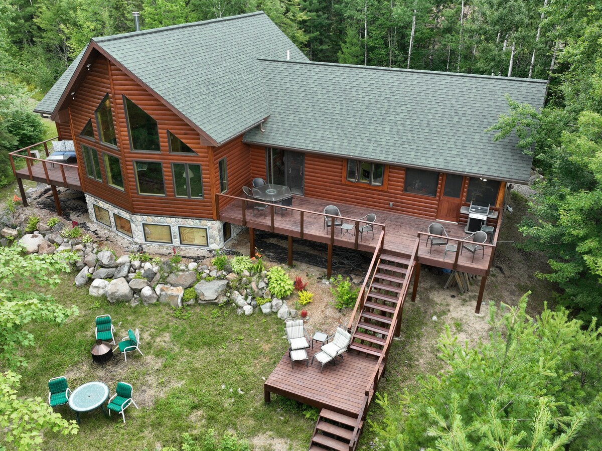 A spacious cabin with a wooden exterior is surrounded by trees. A large deck features multiple seating areas, including chairs and tables. A staircase leads from the deck to the ground level, which is adorned with natural landscaping and two fire pits.