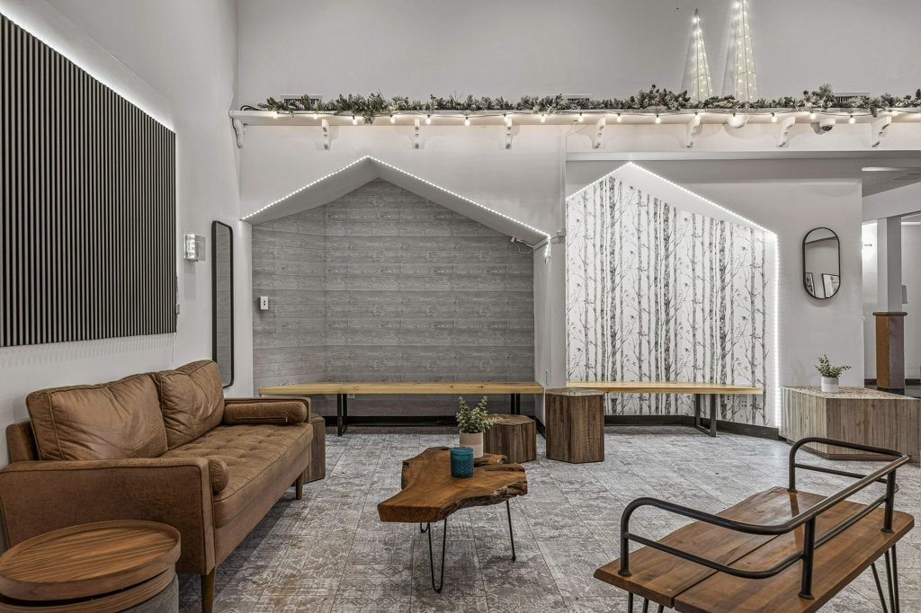 The shared lobby area features minimalist decor with a cozy brown sofa, wooden coffee tables, and benches. Natural light floods the space, highlighting the textured walls and subtle greenery. Decorative wall features include a nature-themed mural and modern lighting fixtures above.
