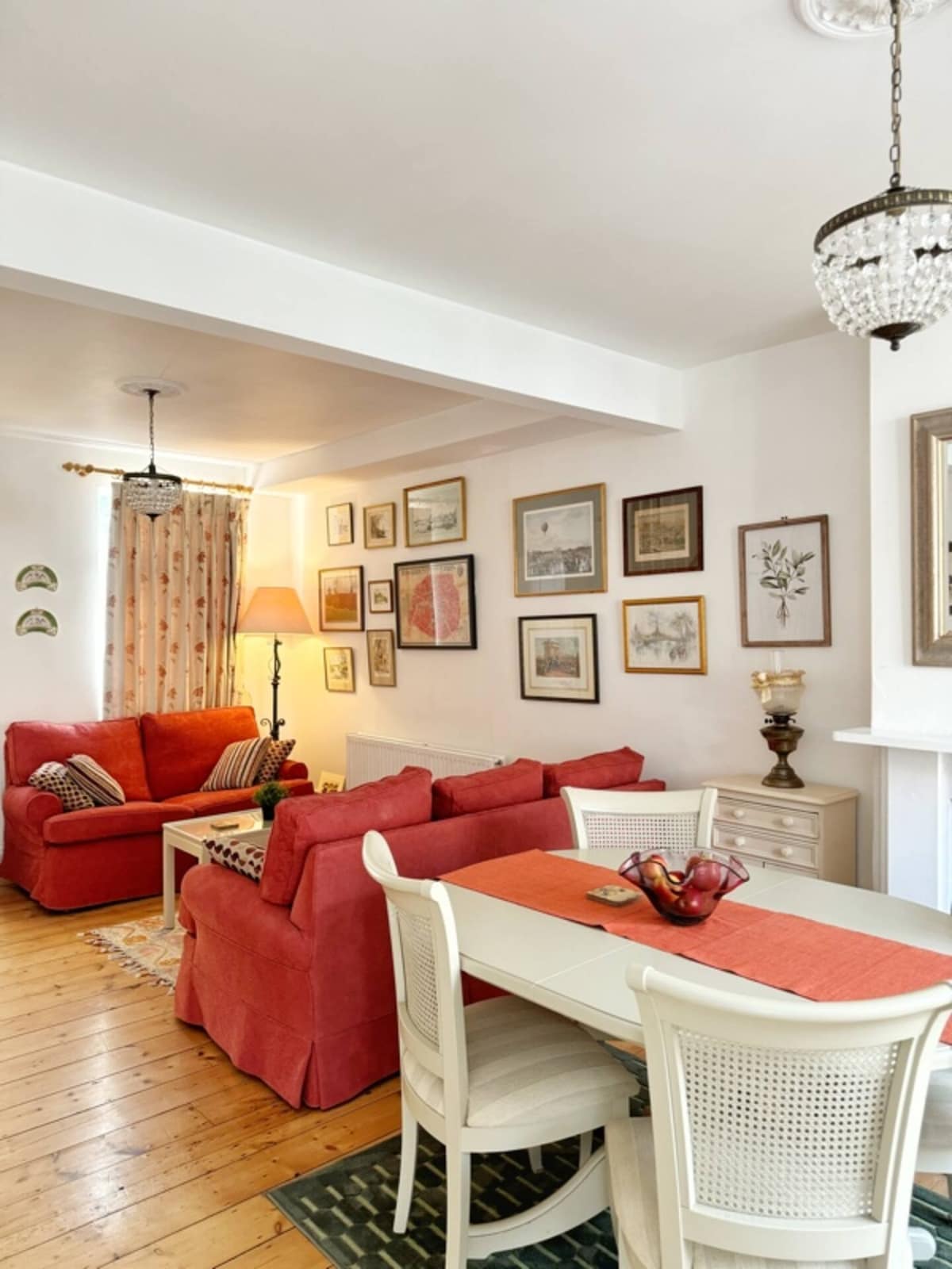 The open lounge and dining area features a cozy layout with red upholstered sofas and a light wooden dining table. Walls are adorned with a variety of framed artwork, contributing to a vintage aesthetic. A warm glow is provided by a hanging light fixture.