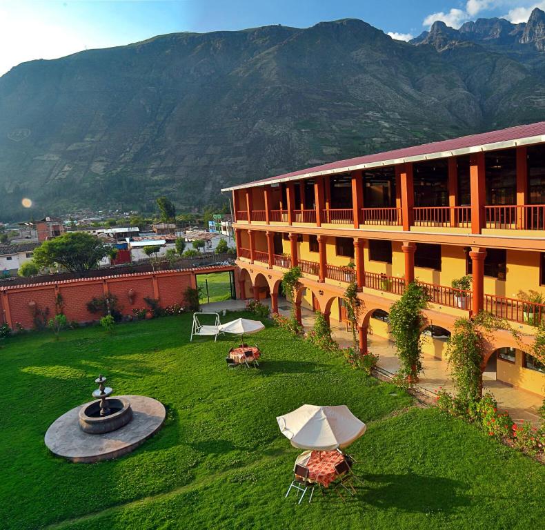 The outdoor area features a lush green lawn, complemented by a central fountain and shaded seating areas. The two-story building, with a spacious balcony, overlooks the picturesque landscape of surrounding mountains and local town activities.