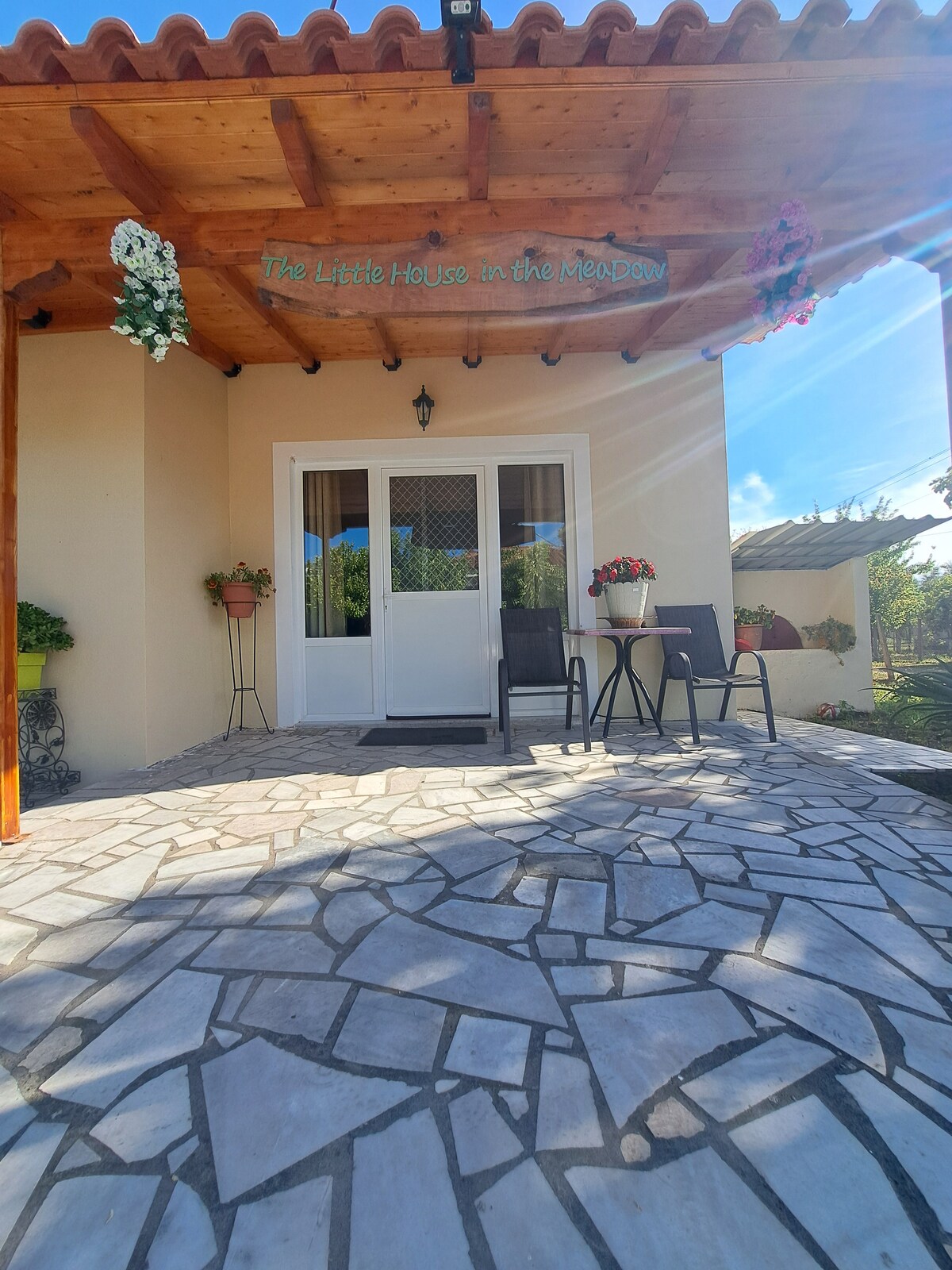 The entrance of the house features an inviting patio with two metal chairs and a small table, surrounded by potted flowers. A welcoming sign hangs above the door, and natural light filters through the open entryway under a wooden roof.