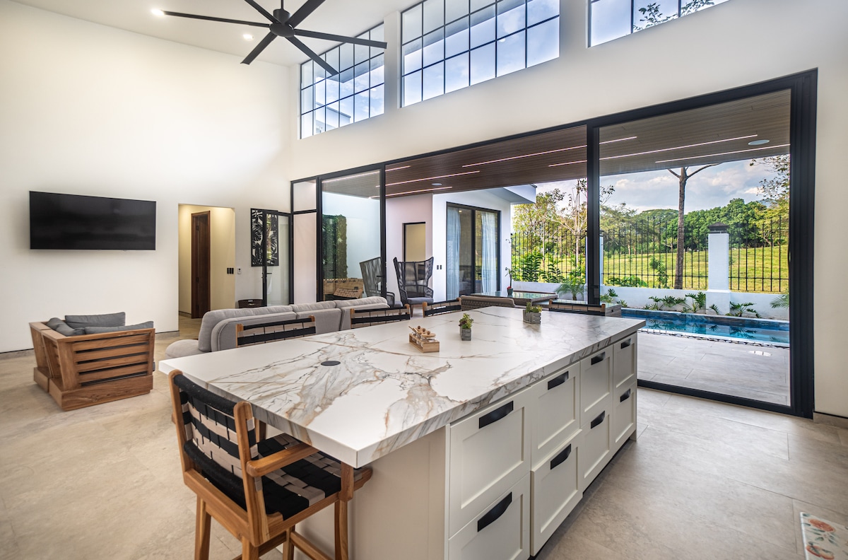 A modern kitchen area features a spacious white island with a marbled countertop, complemented by wooden bar stools. Large glass doors provide a view of the outdoor space, which includes a pool and natural surroundings, while bright natural light illuminates the open layout.