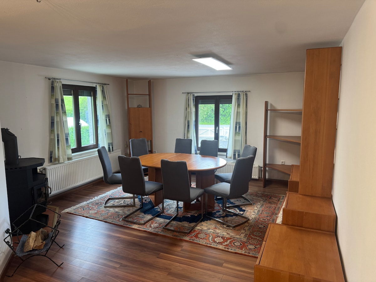 A spacious dining area is equipped with a circular wooden table surrounded by eight grey chairs. Natural light filters through large windows, and two side shelves are positioned against the walls. A traditional rug adds warmth to the hardwood floor, while a black stove stands in one corner.