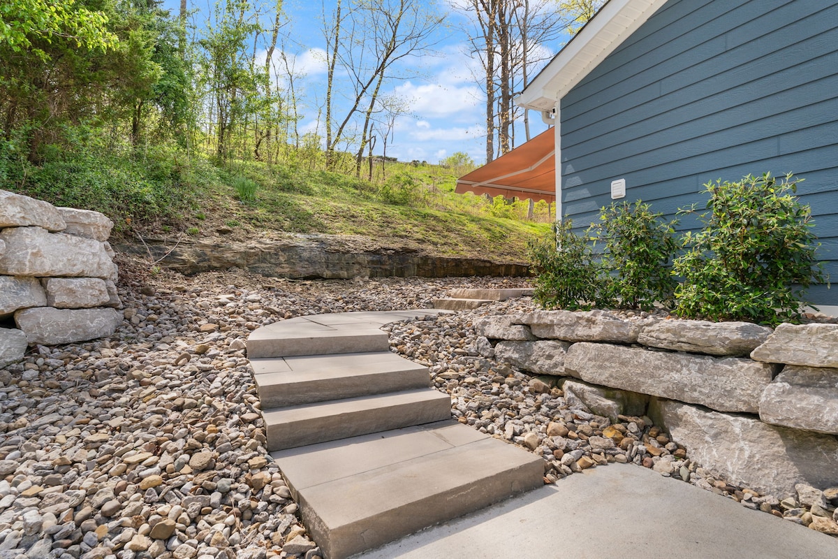 A stone pathway leads to the entrance of the cottage, bordered by carefully arranged rocks and low shrubs. Lush greenery and trees are visible in the background, suggesting a natural and serene environment.