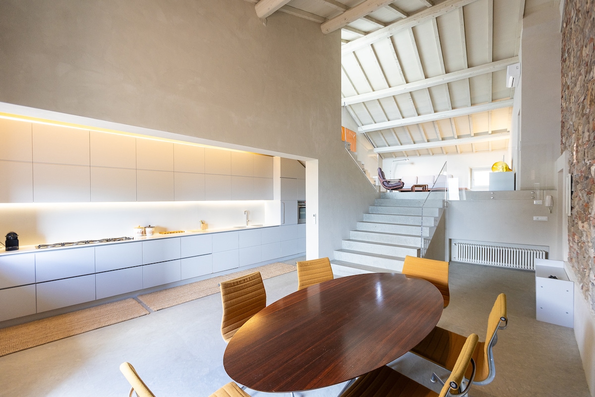 The open-plan dining area features a round wooden table surrounded by six wooden chairs. A sleek kitchen is visible with minimalist cabinetry and modern appliances. Soft lighting illuminates the space, highlighting the high ceilings and natural materials of the walls.