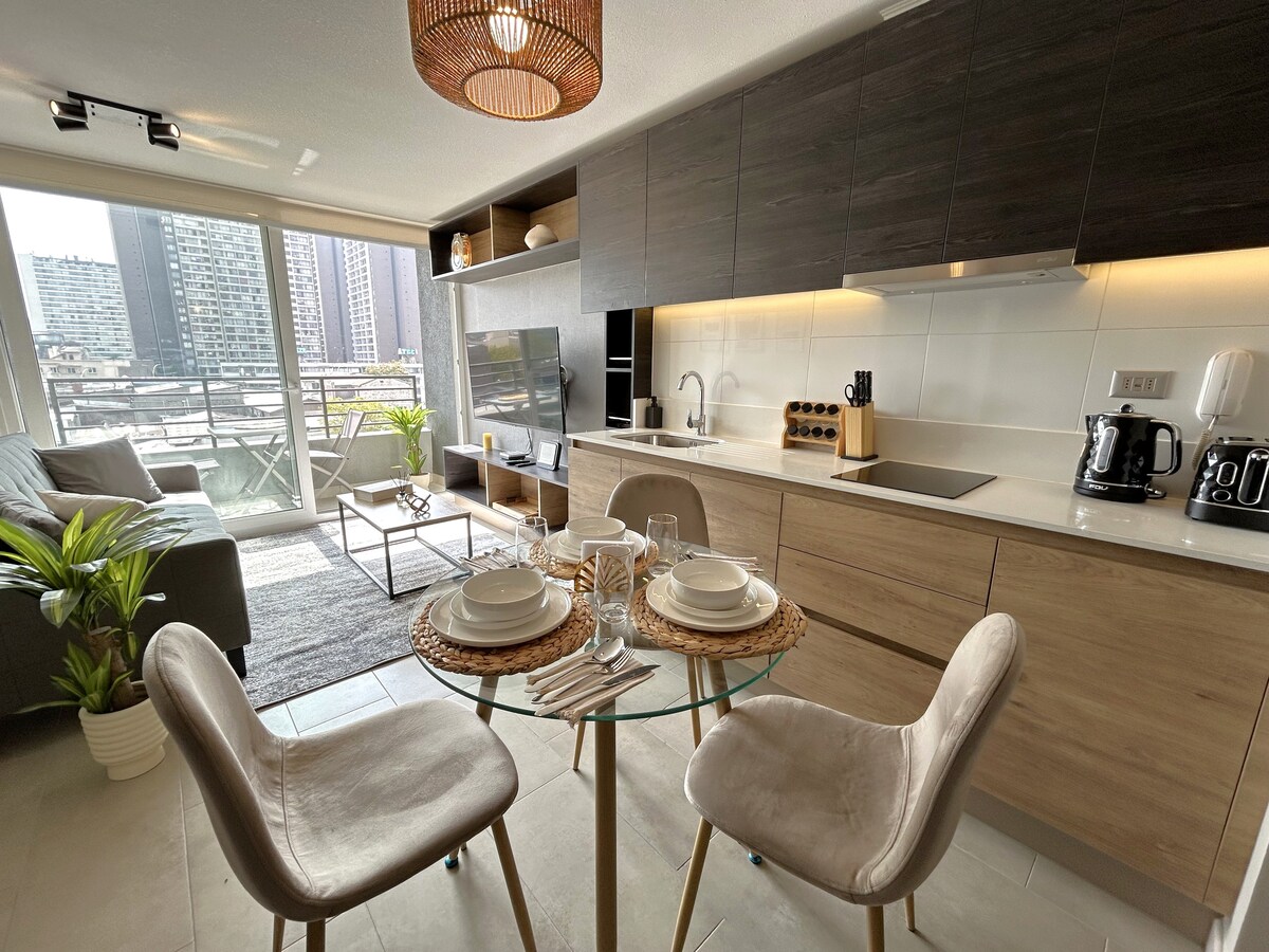 A modern kitchen and dining area are presented, featuring a circular glass table set for two. A stylish couch and coffee table are visible in the background, with large windows revealing views of the city skyline. Natural light enhances the warm wooden tones of the cabinetry.