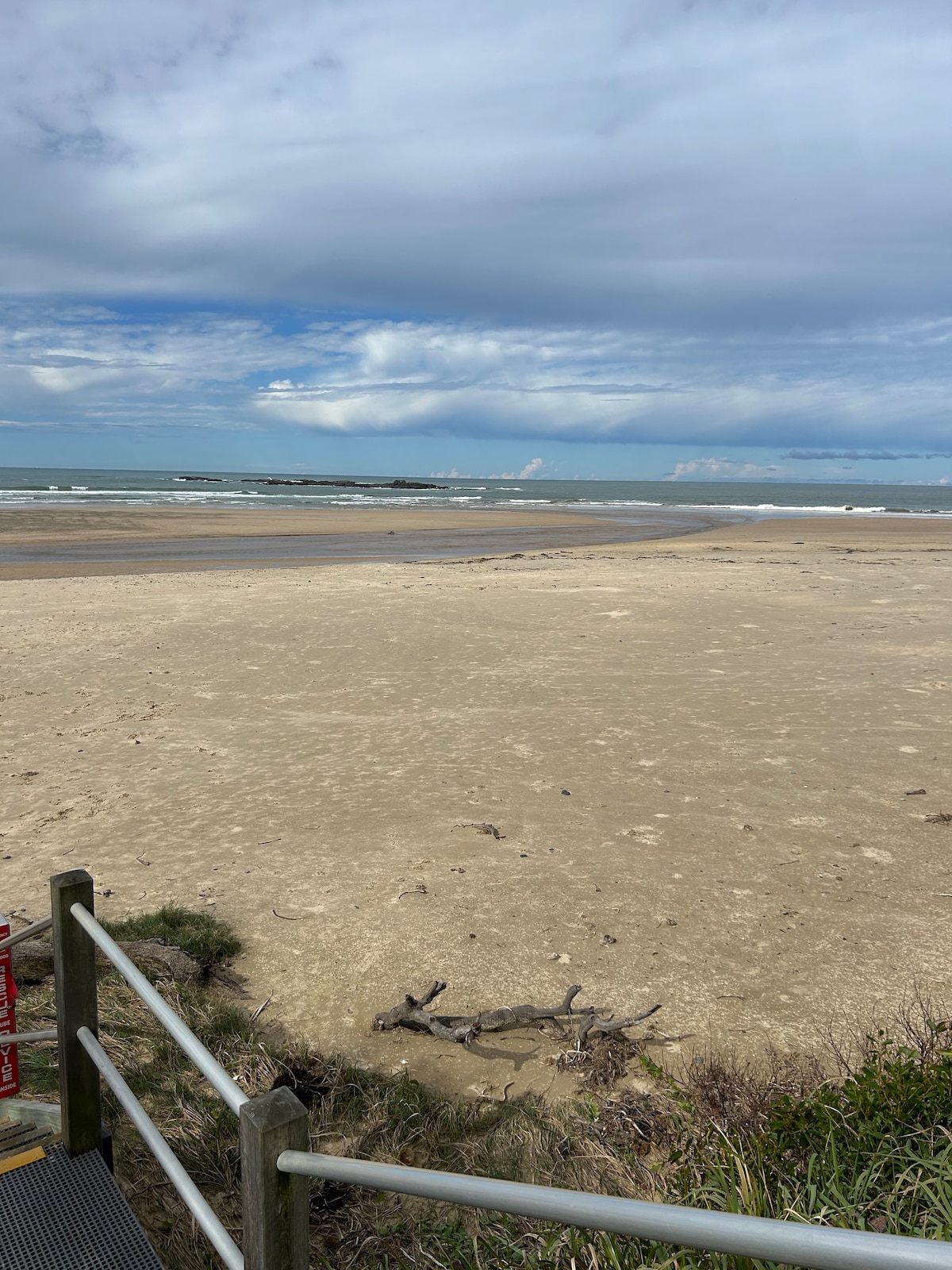 A stretch of sandy beach is visible, bordered by gentle waves lapping at the shore. The sky is filled with soft clouds, creating a calm atmosphere. Dune vegetation is present in the foreground, adding a natural touch to the coastal scene.