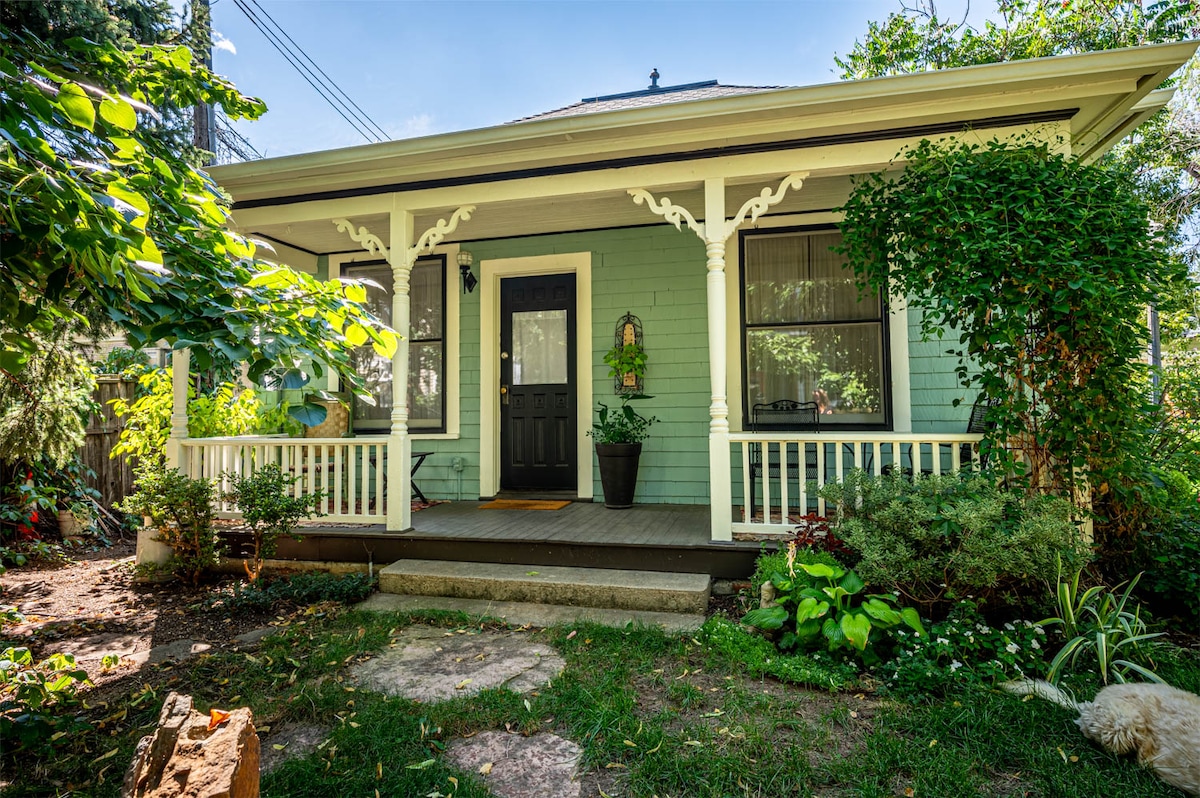 A charming cottage exterior is framed by lush greenery and vibrant gardens. The inviting porch is adorned with decorative woodwork, and a potted flower greets guests by the door. Natural light falls on the peaceful entryway, creating a serene atmosphere.