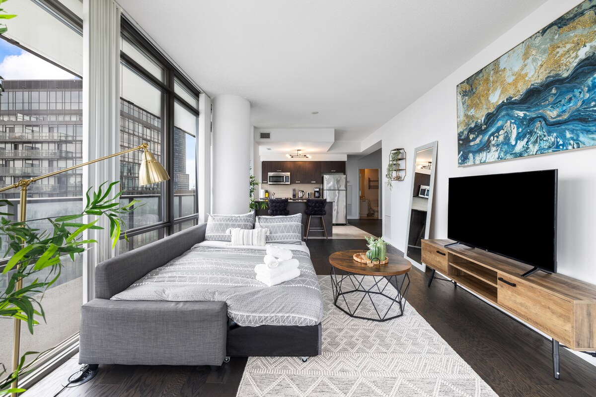 A modern living area showcases a cozy gray sofa that converts into a bed. A circular wooden coffee table is situated on a patterned rug. Large windows allow natural light to fill the space, while a flat-screen TV is mounted against the wall.