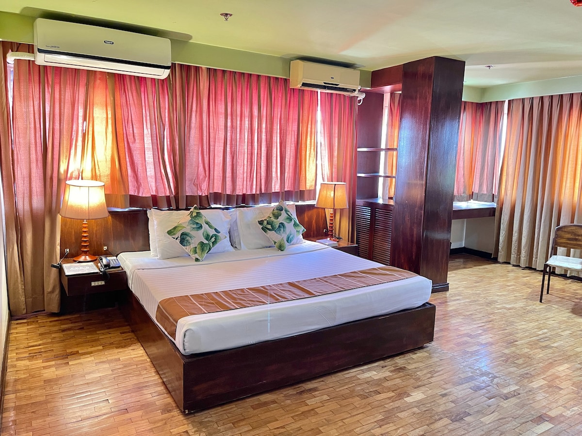 A spacious sleeping area is highlighted by a large bed with decorative pillows. Soft lighting from two bedside lamps creates a warm ambiance. Window curtains in soft pink filter natural light, while wooden flooring adds a natural element to the room's design.