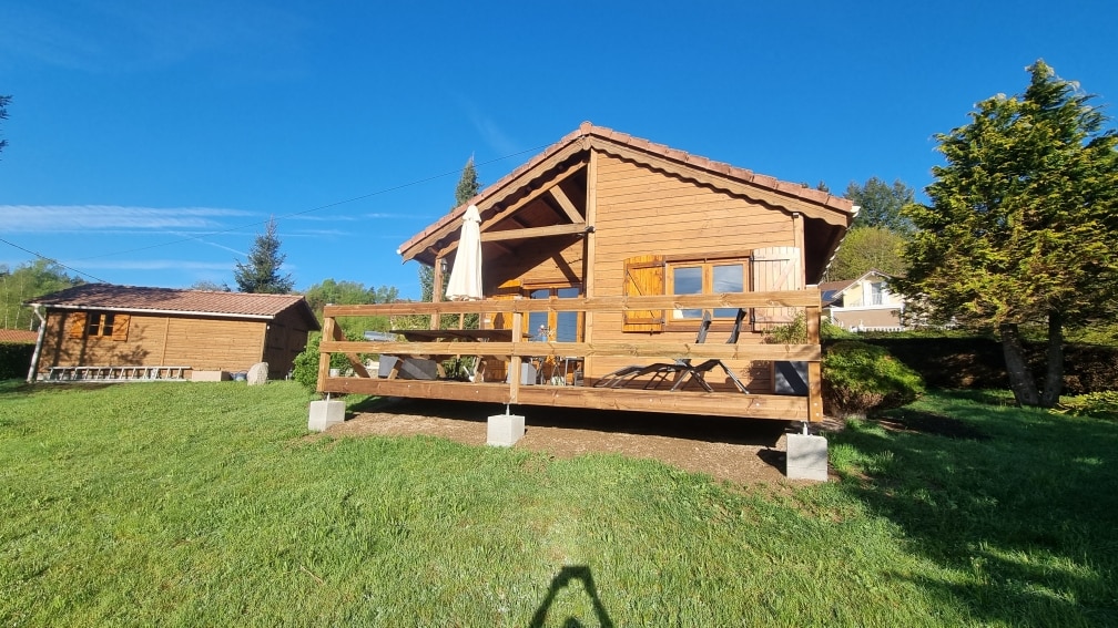 A charming wooden chalet is set in a green landscape under a clear blue sky. The structure features a spacious front deck with comfortable seating and a sun umbrella, complemented by vibrant greenery surrounding the property.