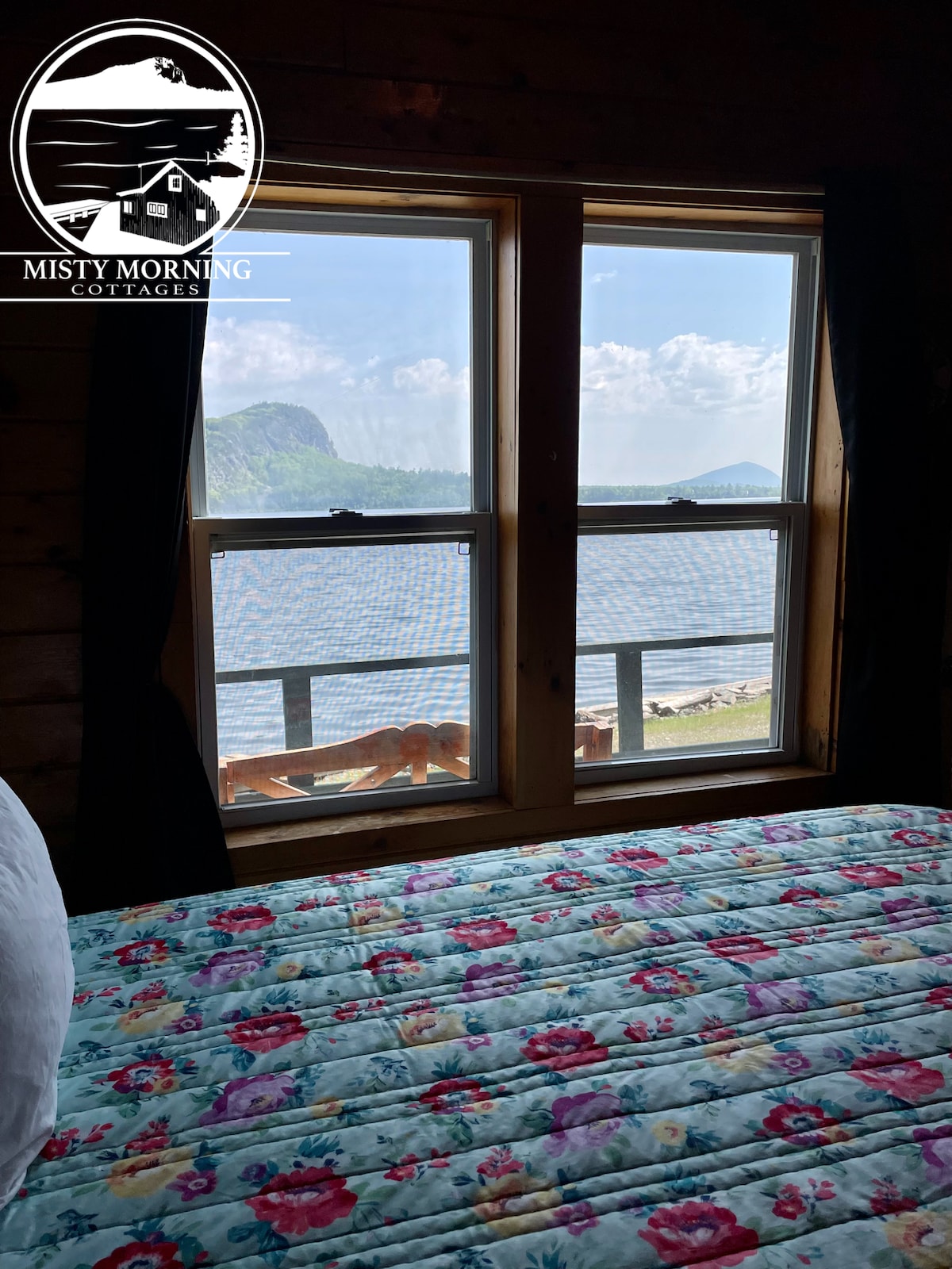 A view from inside the cottage reveals a window overlooking Moosehead Lake, framed by two dark curtains. A spacious bed with a floral-patterned quilt is positioned nearby, inviting natural light and offering a glimpse of Mt. Kineo and the serene landscape beyond.
