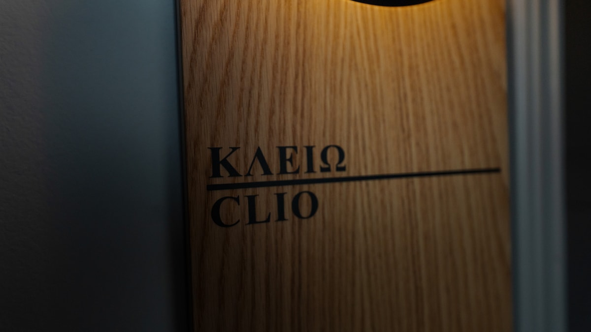 A wooden door sign displays the names 'ΚΛΕΙΩ' and 'CLIO' in contrasting black lettering. Soft lighting highlights the texture of the wood, while the smooth surface blends harmoniously with the surrounding wall.