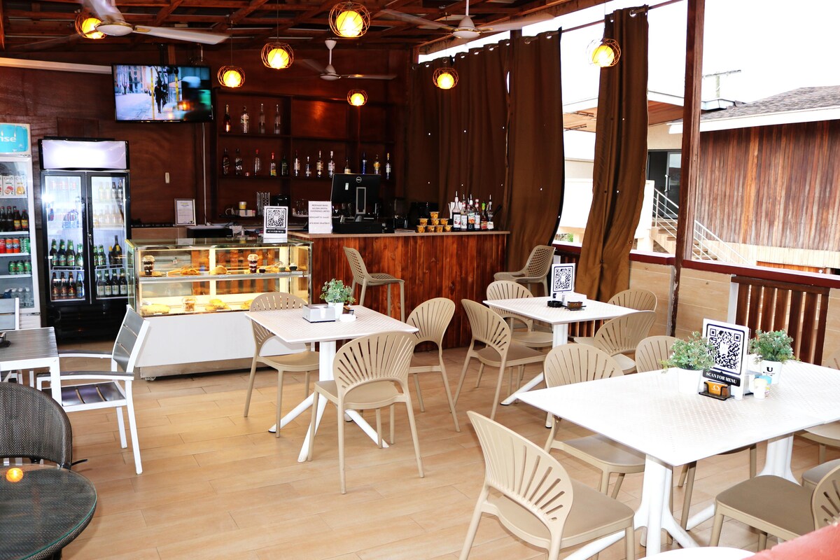 An inviting bar area features a wooden counter with bar stools and a display of beverages. Multiple tables and chairs provide seating for guests. Soft lighting and decorative plants enhance the space, while large windows allow natural light to fill the area.