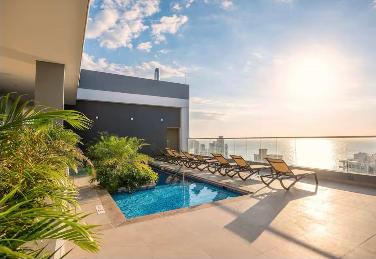 A rooftop pool area features a serene setting with a small pool surrounded by lounging chairs. Lush greenery adds a natural touch, and expansive views of the ocean are visible in the distance, illuminated by soft sunlight.