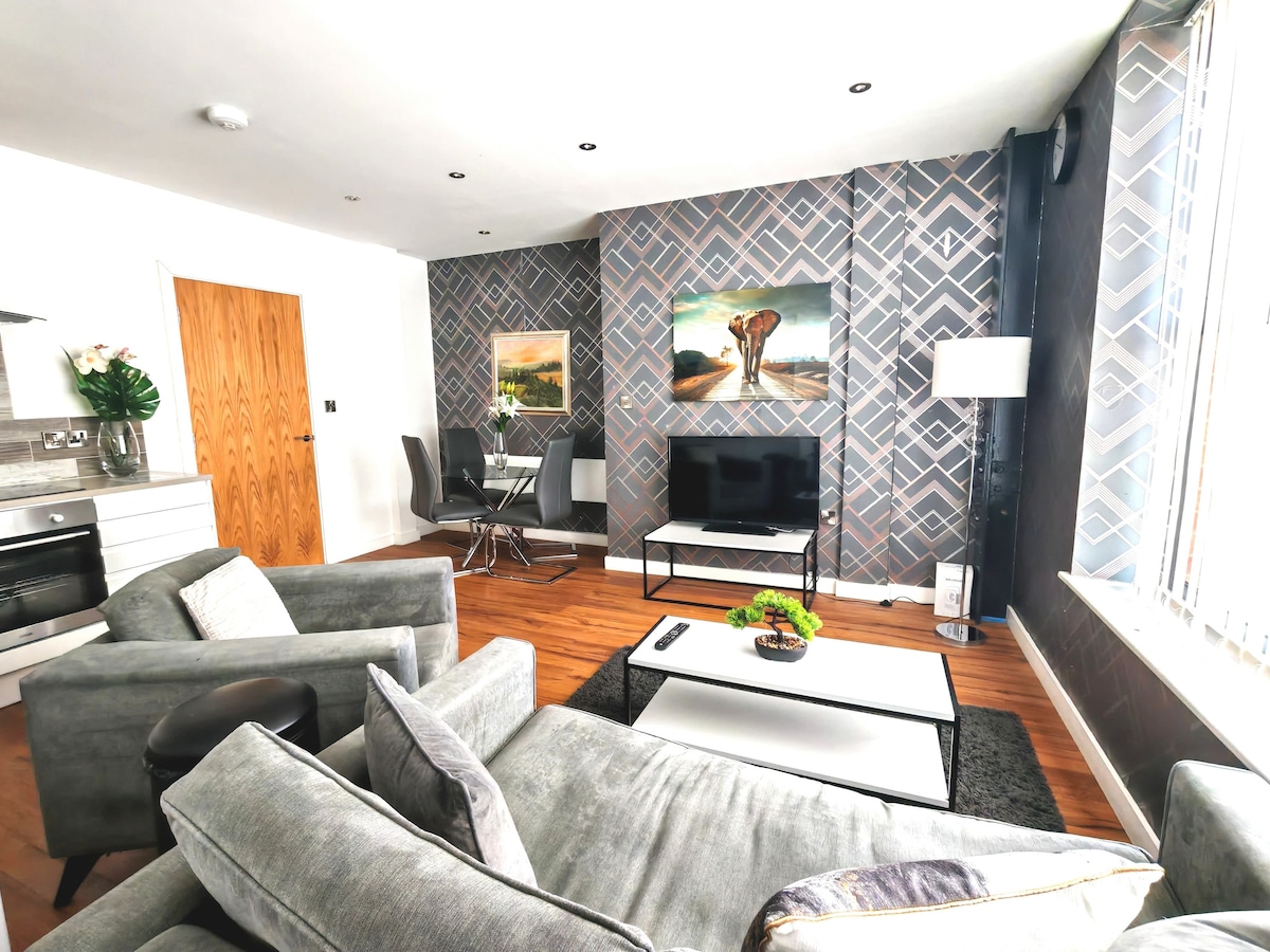 A modern living area features a grey sectional sofa and a stylish coffee table. A flat-screen television is positioned against an accent wall with geometric patterns. A dining table for four is visible, complemented by a large window allowing natural light into the space.
