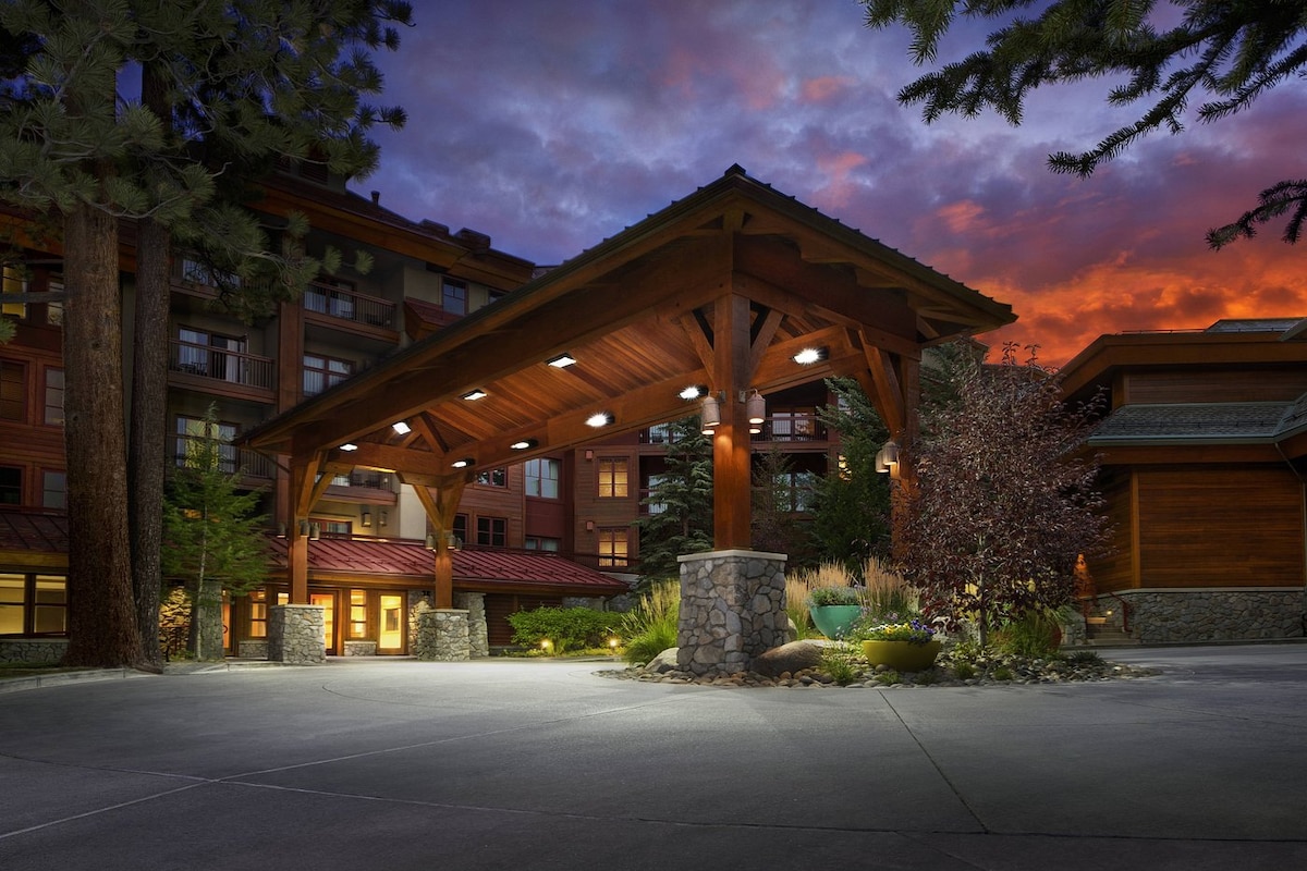 The entrance features a large wooden portico with multiple lights illuminating the area. Surrounding greenery and decorative stone elements enhance the natural ambiance. The backdrop includes a building structure with rustic architecture, under a colorful sky at dusk.