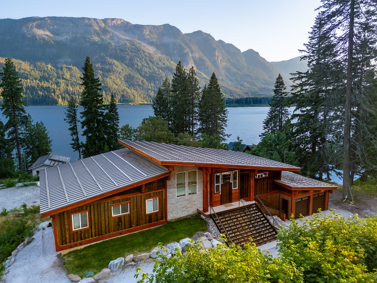 The cabin is situated on the shoreline of Lake Wenatchee, surrounded by tall evergreens and stunning mountain views. The modern architecture features large windows and a spacious deck. A pathway leading to the entrance is visible, enhancing the inviting exterior of the home.