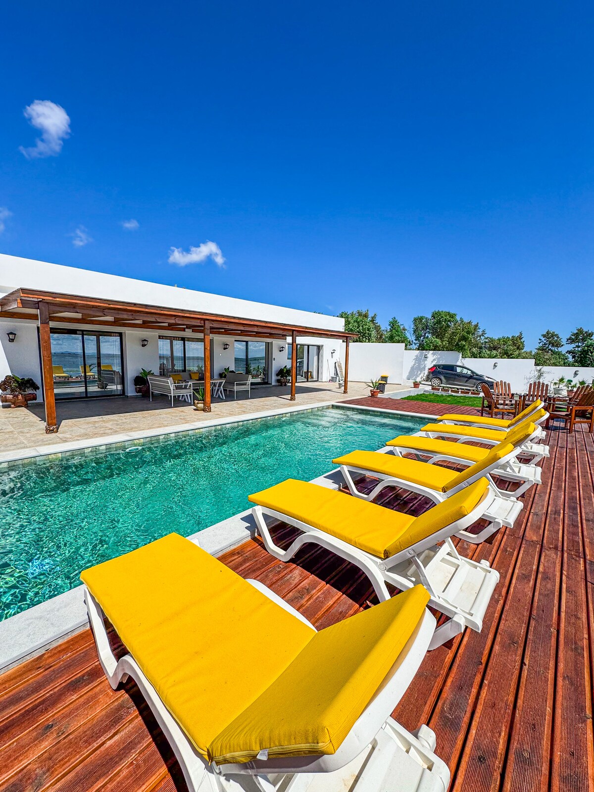 A row of yellow lounge chairs is positioned beside a clear swimming pool, bordered by wooden decking. The pool reflects the vibrant blue sky and is flanked by a modern structure with large glass doors, providing views of the surrounding landscape.