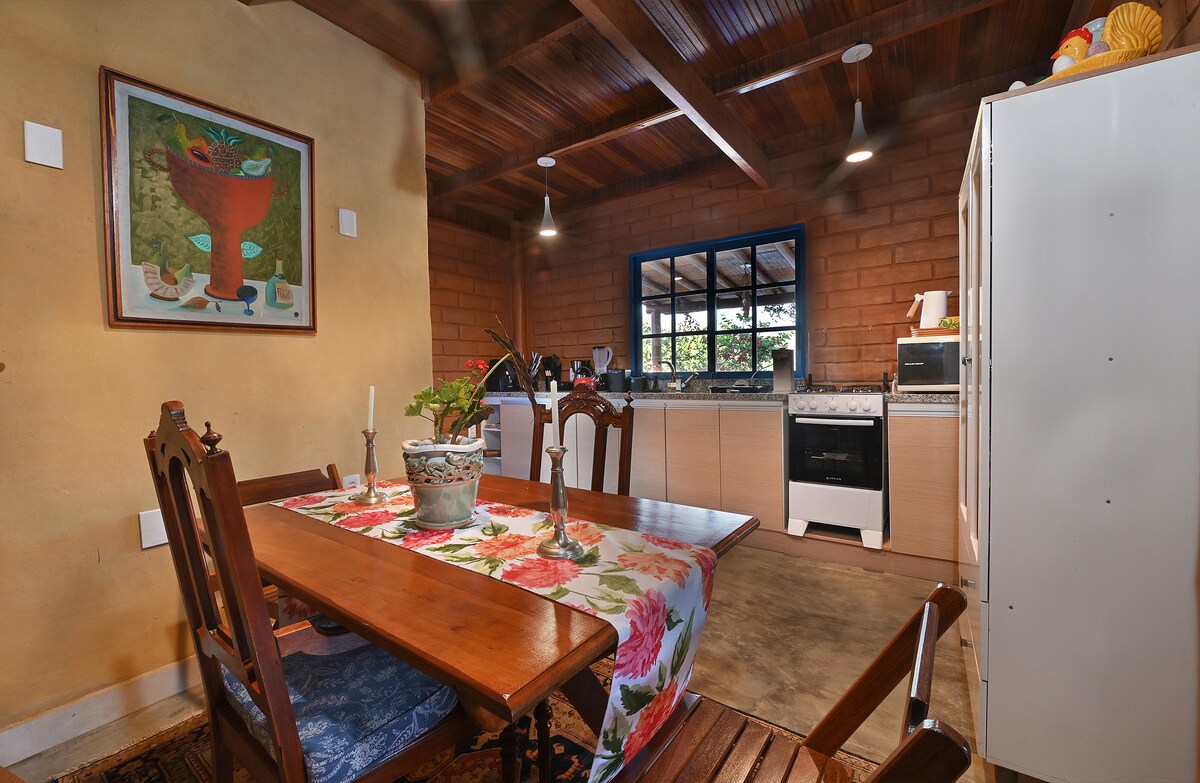 A rustic kitchen area features wooden beams and a charming dining table set for four, adorned with a floral tablecloth. A range of appliances, including a stove and refrigerator, are positioned against a wall with natural stone accents. Large windows allow natural light to illuminate the space.