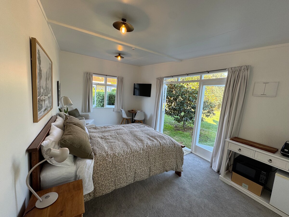 A self-contained room features a spacious bed with a light-toned coverlet and multiple cushions. A SmartTV is mounted on the wall opposite a small dining table. Large windows allow natural light to fill the space and provide views of the garden outside.