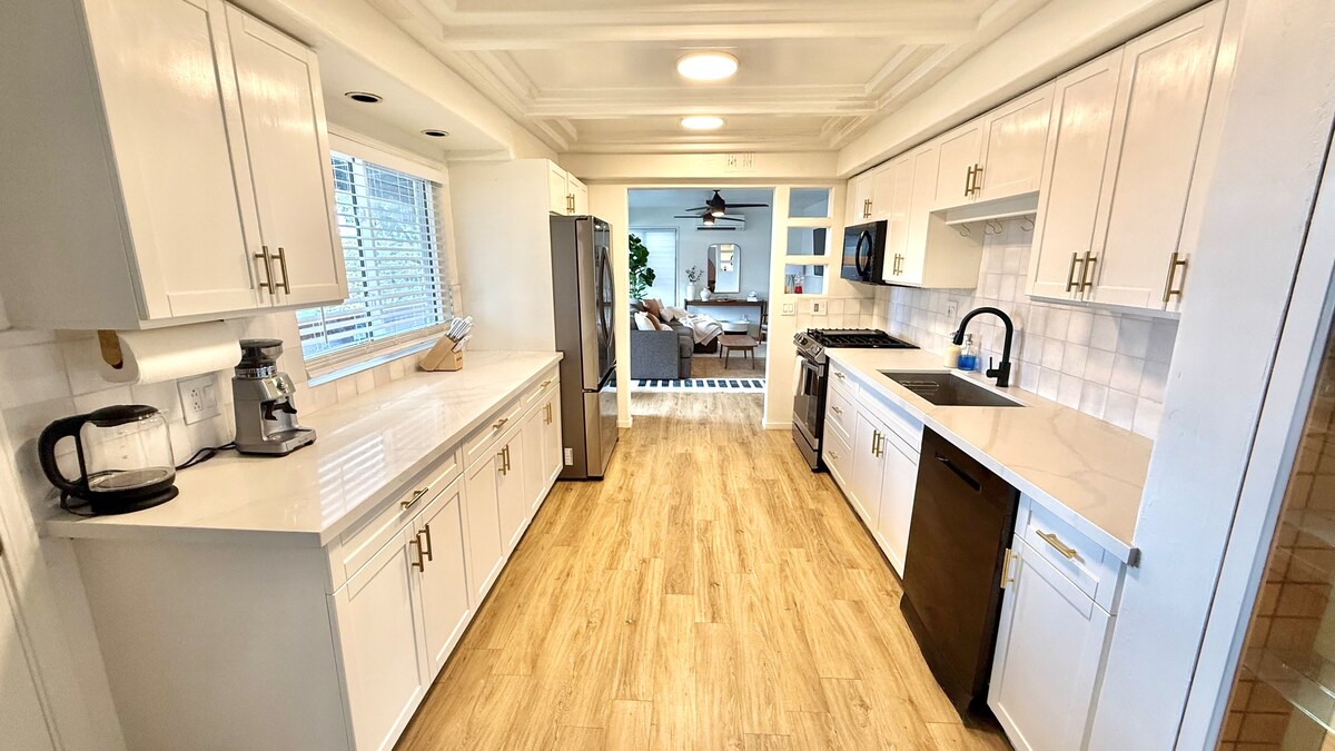 A modern kitchen features white cabinetry and countertops, with stainless steel appliances including a refrigerator and oven. Natural light spills in through a window, illuminating the spacious layout with ample counter space and a back door leading to another area of the home.