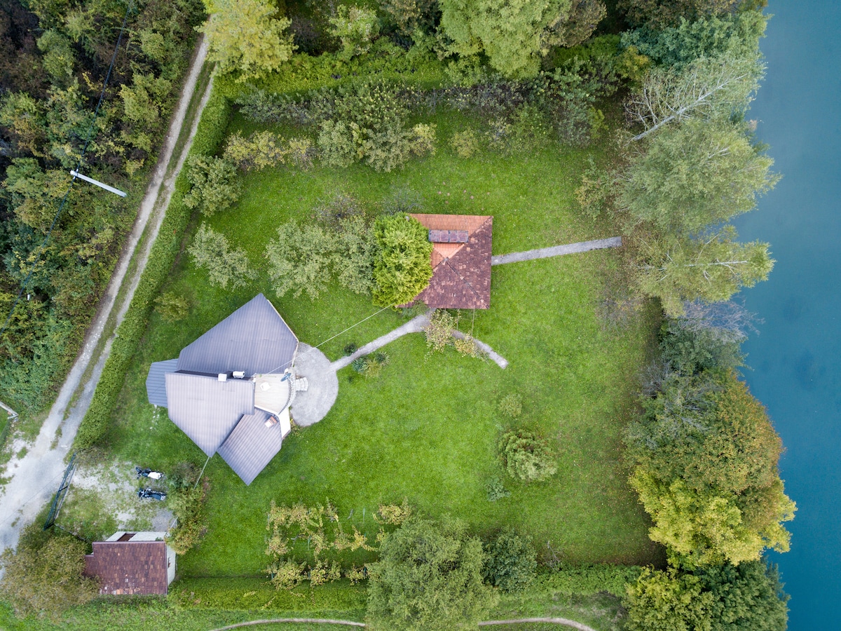An aerial view captures a spacious garden surrounded by lush greenery, featuring a winding path leading to a cozy riverside retreat. The house's distinct roof design is seen alongside a separate structure, with the tranquil river visible at the edge of the property.