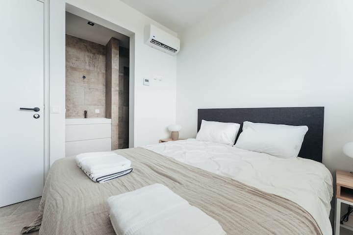 Dieses ruhige Schlafzimmer mit eigenem Bad verbindet modernen Minimalismus mit gemütlichem Komfort – mit weichen Betten, warmem Licht und einem direkten Blick in ein elegantes, steinverkleidetes Badezimmer für nahtlose Entspannung.
