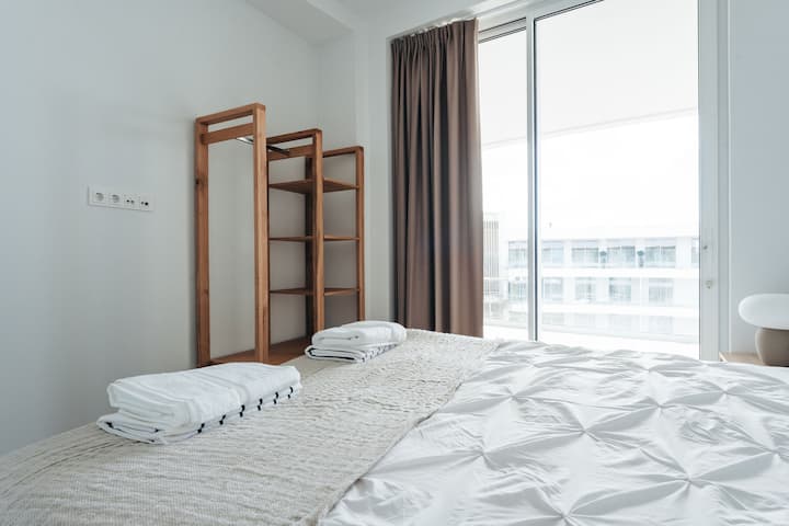Dieses helle und luftige Schlafzimmer kombiniert weiche Texturen mit minimalistischem Design – mit einem offenen Kleiderschrank aus warmem Holz, Verdunkelungsvorhängen und Schiebetüren, die sich zu einem sonnendurchfluteten Blick auf die Terrasse öffnen.