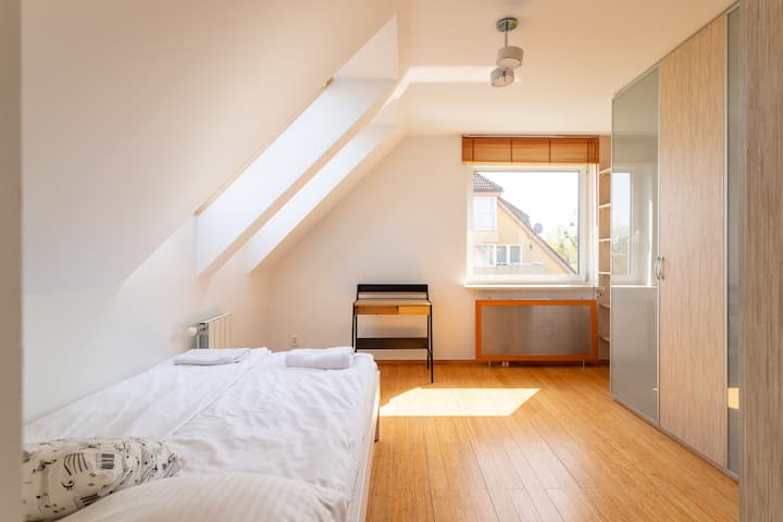 Une chambre confortable avec un lit simple sous un plafond en pente et un bureau près de la fenêtre. La lumière naturelle inonde la pièce à travers une lucarne.