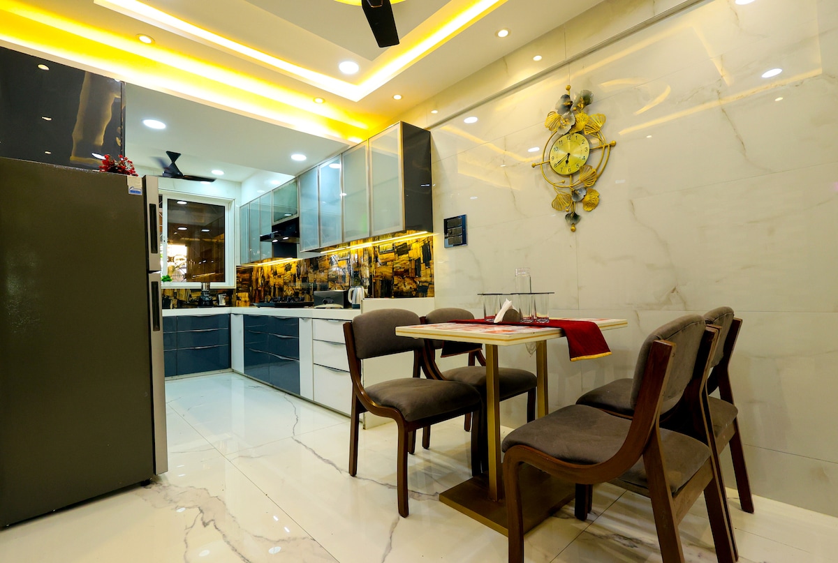 A modern kitchen features sleek cabinetry and ample counter space. A round dining table is surrounded by four comfortable chairs. A fridge stands against the wall, and a vibrant wall clock adds a touch of character to the room. Warm lighting enhances the inviting atmosphere.