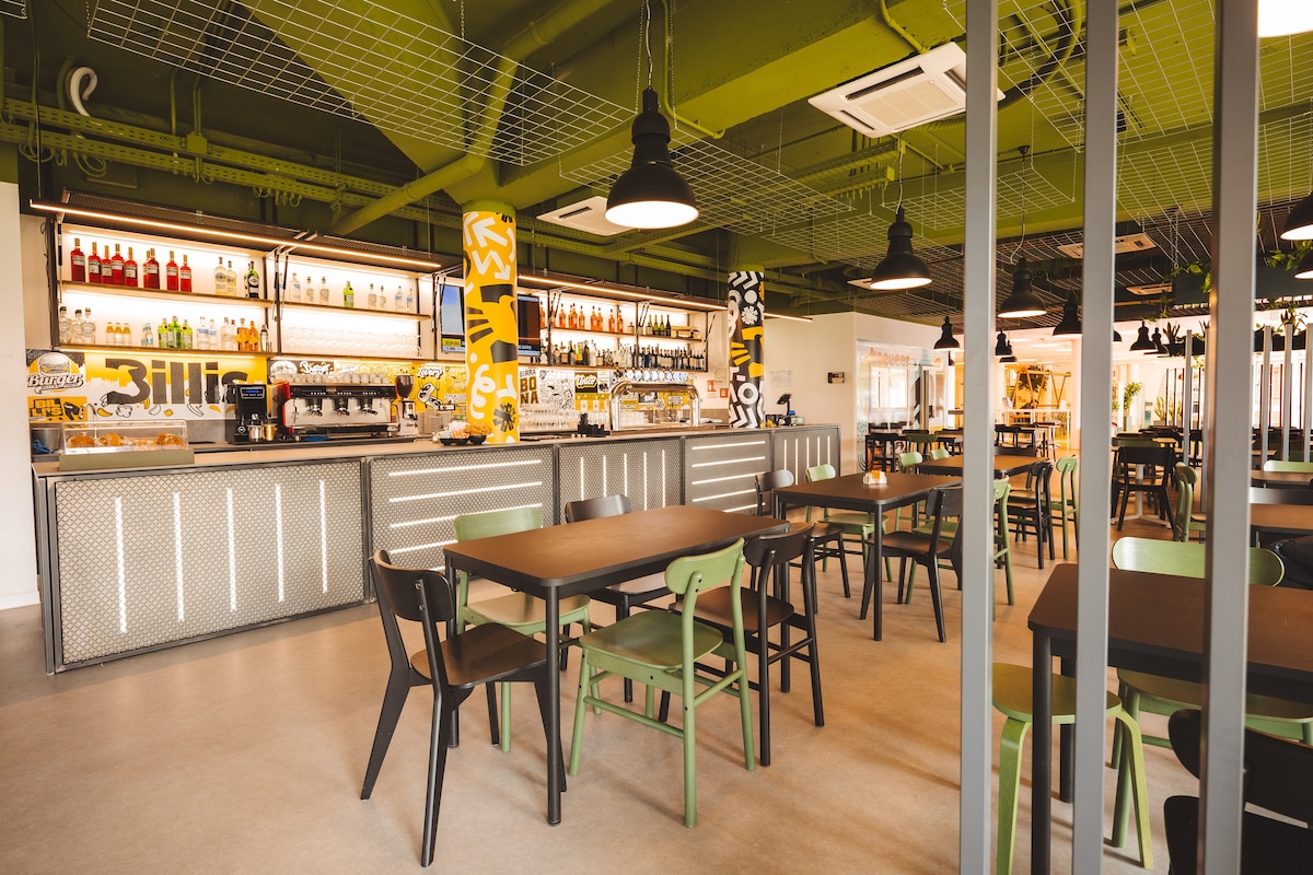 An open dining area features a bar with a variety of spirits displayed. Modern green and black chairs surround dark wooden tables. The ceiling showcases an industrial design, while bright yellow and white graphics add a lively touch to the surroundings.