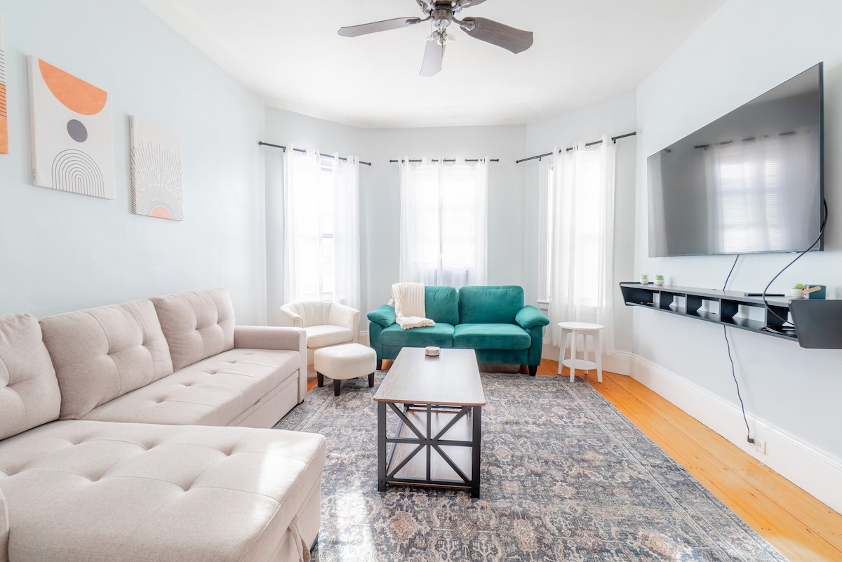 A spacious living room features a light-colored sectional sofa and a green armchair, complemented by a coffee table at the center. Large windows allow ample natural light, with sheer white curtains gently framing the view. A mounted TV and decorative wall art enhance the inviting atmosphere.
