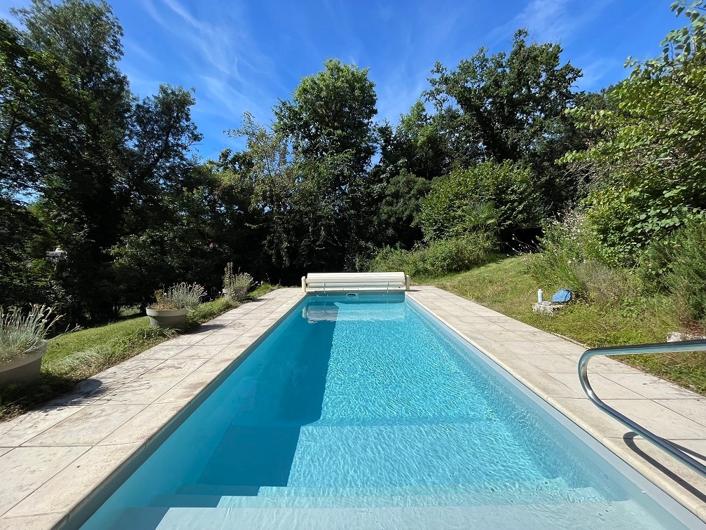 A clear blue swimming pool is centrally positioned, surrounded by a lush garden with trees and shrubs. The smooth stone deck contrasts with the vibrant greenery, and the serene atmosphere is enhanced by the bright blue sky above.