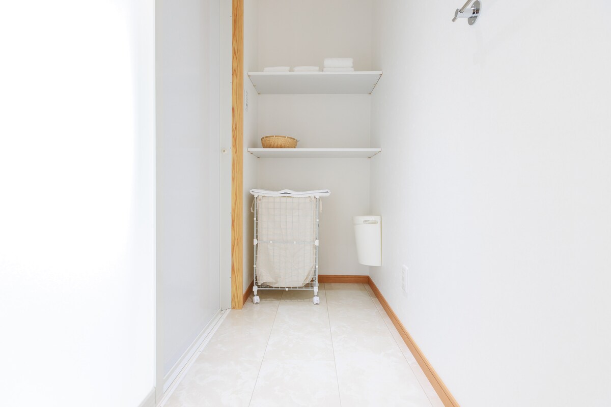 A clean and bright laundry area is displayed, featuring white walls and tiled flooring. Open shelves hold neatly stacked towels, while a wicker basket offers additional storage. A white laundry hamper on wheels is positioned against one wall, contributing to the functional design.