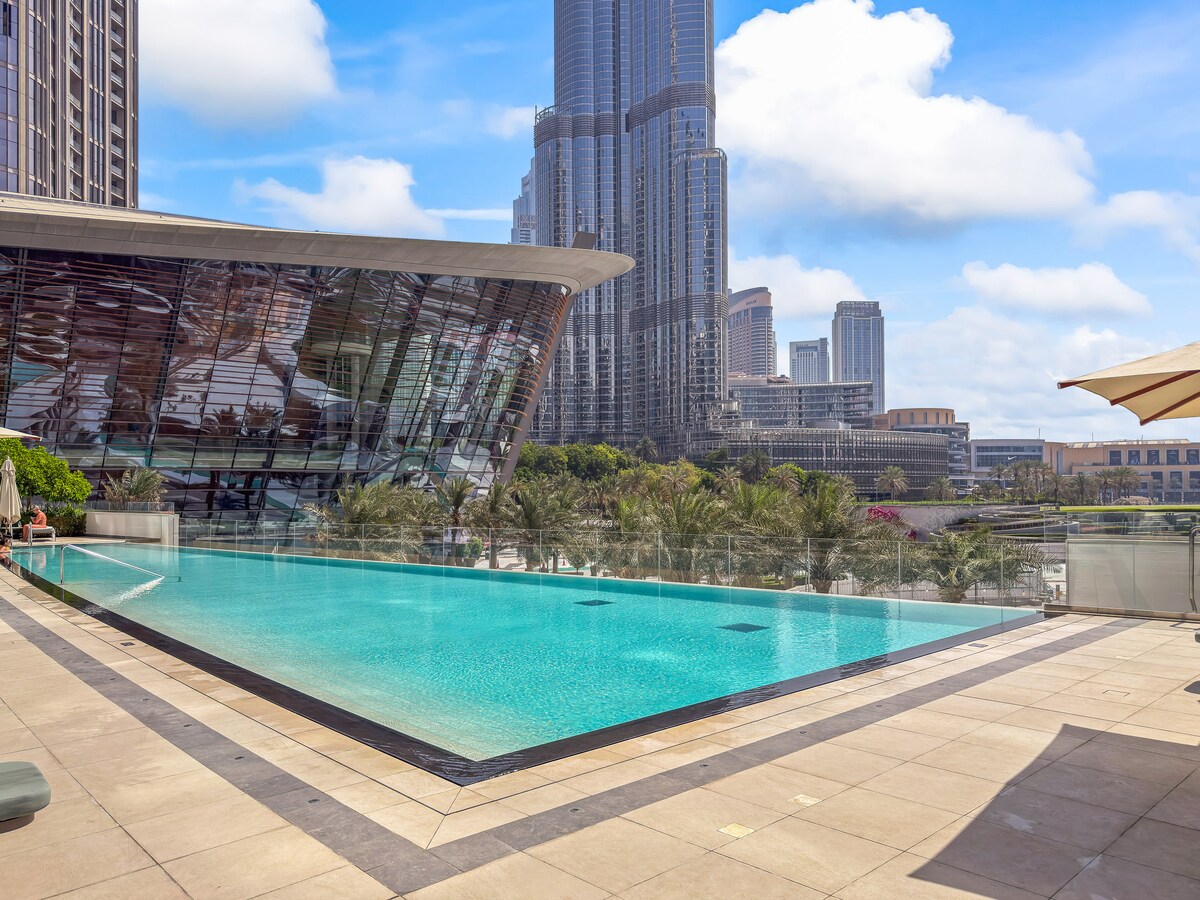 The image showcases an infinity-edge pool surrounded by a landscaped area, with the iconic Burj Khalifa towering in the background. Clear blue skies reflect off the water's surface, while modern architectural elements of the adjacent building create a sleek and inviting atmosphere.