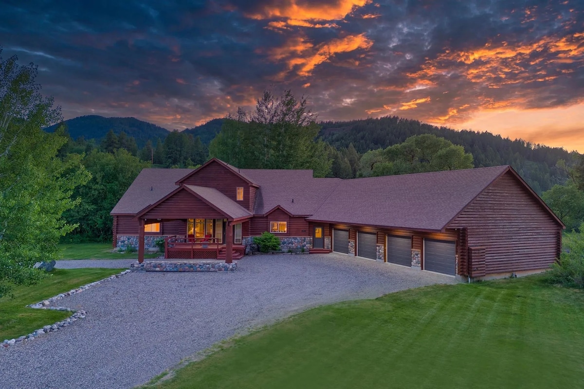 The large wooden structure is set against a stunning sunset sky, with vibrant orange and purple tones. A gravel driveway leads to the front entry, surrounded by grassy areas and trees, with mountain silhouettes visible in the background.