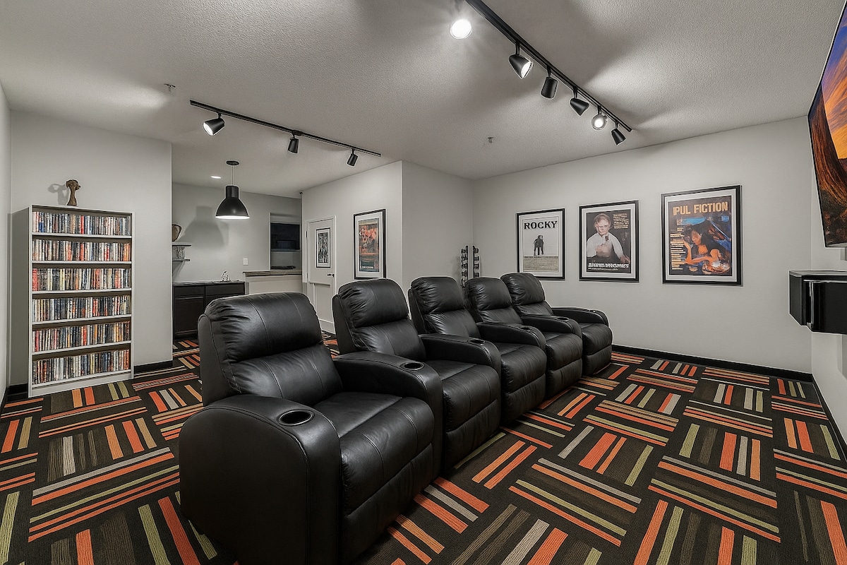 A cozy media room is equipped with five black reclining chairs, each featuring cup holders. Colorful patterned carpeting adds a vibrant touch, while framed movie posters decorate the walls. Bright overhead lighting illuminates the space, enhancing its welcoming ambiance.