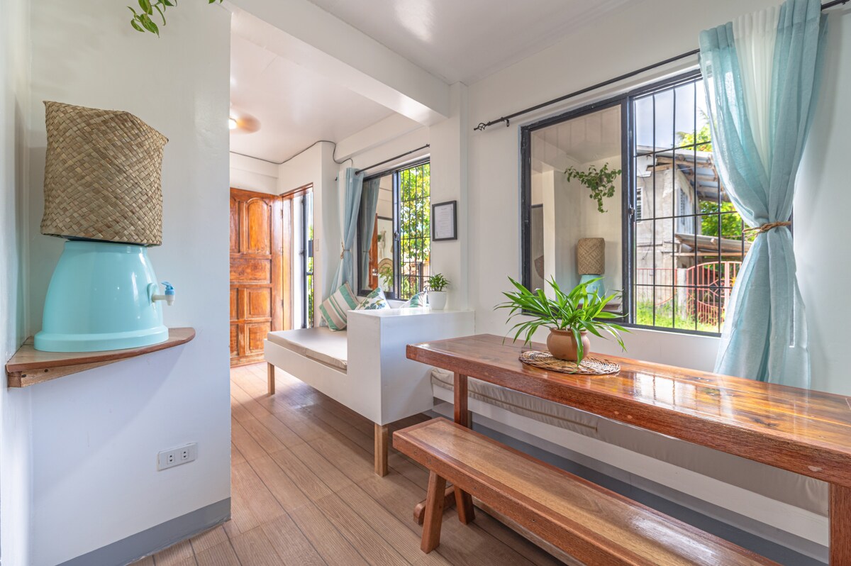 A bright dining area is furnished with a wooden table and benches, accommodating guests comfortably. Light filters through large windows, enhancing the airy atmosphere. A water dispenser is positioned nearby, and a soft seating area features cushions, promoting relaxation. Decor elements include potted plants and wall art.