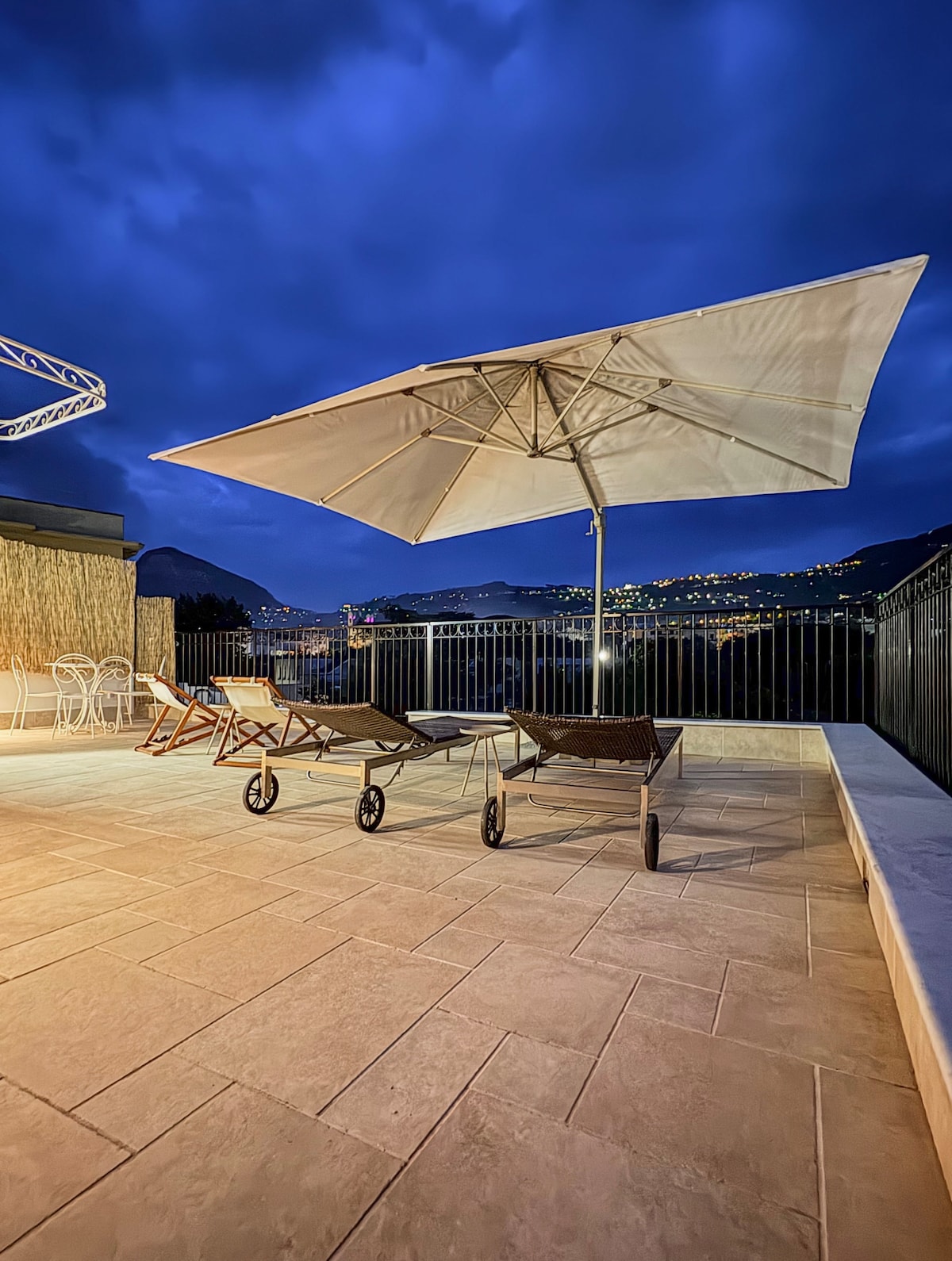 A spacious terrace is seen under a night sky, featuring two lounge chairs and a large umbrella. The backdrop includes distant lights from the surrounding hills, creating an atmospheric setting for relaxation.
