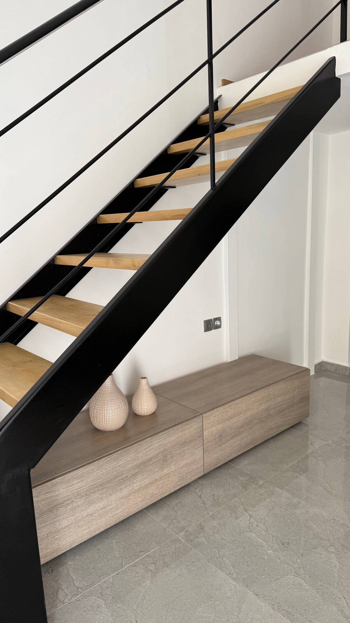 A modern staircase is depicted with wooden steps contrasting against a black metal frame. Below, a sleek wooden console table holds two decorative vases. The floor features a polished stone surface, enhancing the contemporary feel of the entryway.