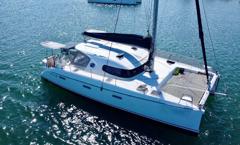 The catamaran is anchored in calm waters, featuring a modern design with a spacious deck. Sunlight reflects off the surface of the water, highlighting the sleek lines of the vessel. A shaded area on the deck provides a comfortable space for relaxation.