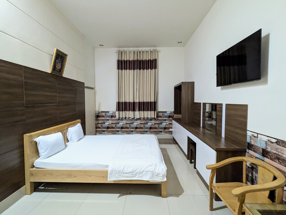 A modern room is displayed with a spacious bed against a dark accent wall, complemented by light-colored bedding. A wooden chair is positioned next to a minimalist desk, while a television is mounted above a sleek storage unit. Soft natural light filters through the window covered with curtains.