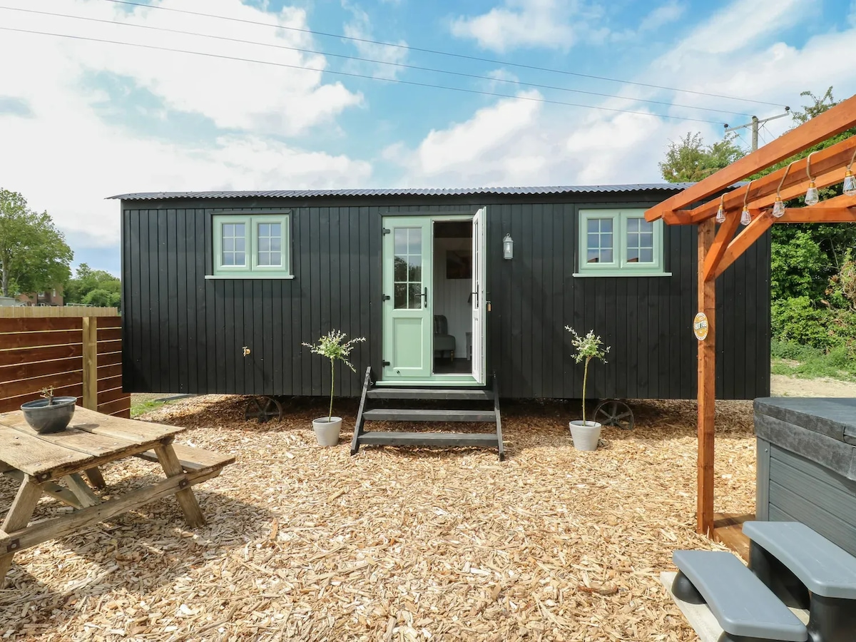 The exterior of the shepherd hut is displayed, featuring a dark wood finish with light green accents. A welcoming entrance is flanked by two small windows, revealing a compact outdoor area with picnic seating and potted plants. The hot tub is partially visible to the right.