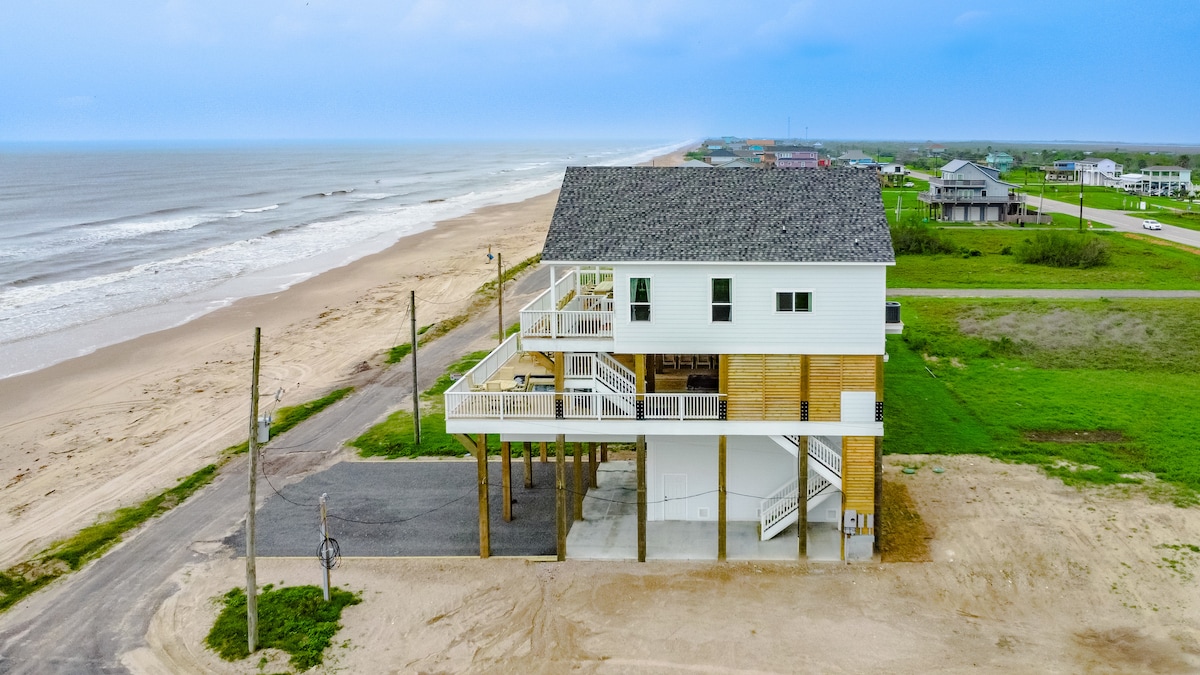 An aerial view of a modern coastal beach house elevated on stilts, situated beside a sandy beach. The property features multiple levels with expansive balconies, providing unobstructed views of the ocean. Nearby, the shoreline meets the waves, with a green expanse indicating a serene backdrop.